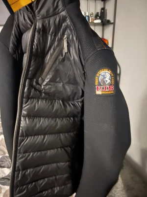 Svart pufferjacka från Parajumpers - Svart pufferjacka från Parajumpers med huva och coolt patch på ärmen. Jackan har quiltad framsida och rygg, dragkedja framtill och praktiska fickor med orange detaljer. Materialmix av polyester och syntet, perfekt för kyliga dagar.