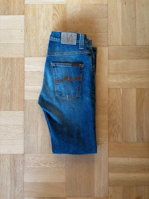 Blå jeans från Nudie Jeans - Säljer ett par klassiska blå jeans från Nudie Jeans med snygga slitningar och kontrastsömmar på bakfickorna. Modellen har rak passform och normal midja, tillverkade i slitstarkt bomullsmaterial. Perfekt för dig som gillar en tidlös och cool look.