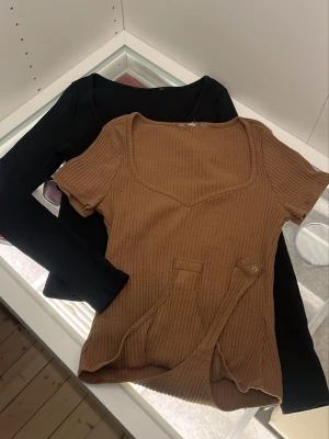 Ribbad brun och svart topp - Två ribbade toppar säljes, en brun kortärmad med v-ringning och dekorativa knappar framtill, samt en svart långärmad med rund hals. Båda har en slim passform och är gjorda i mjukt, stretchigt material som känns skönt mot huden.