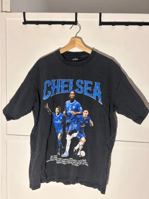 Chelsea vintage t-shirt XL från Golazo - Säljer en svart vintage Chelsea t-shirt från Golazo i storlek XL. Stor tryck fram och bak med Chelsea-spelare och texten 'Winners 2005'. Skön bomullskänsla och loose fit, perfekt för dig som älskar fotboll och retrostil.