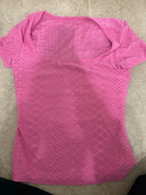 Rosa spetsblus med kort ärm - Säljer en söt rosa blus med korta ärmar och spetsdetaljer i ett geometriskt mönster. Toppen har en fyrkantig halsringning och är tillverkad i ett lätt och luftigt material som passar perfekt till varmare dagar.