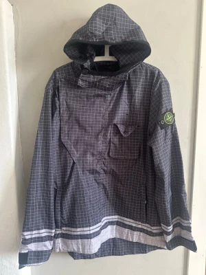 Grå rutig vindjacka Stone Island XL - Snygg grå vindjacka från Stone Island med rutigt mönster och stor huva. Jackan har vita och svarta detaljer längst ner, bröstficka med knapp och klassisk Stone Island patch på ärmen. Perfekt för dig som vill ha streetwear-vibe och stilren look.