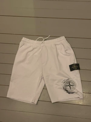 Vita Stone Island shorts med snörning - Säljer ett par vita shorts från Stone Island med elastisk midja och snörning. De har en stor logga tryckt på ena benet och en svart patch med broderad logga på sidan. Materialet är mjuk bomull och bakfickan har dragkedja. Perfekta för chill dagar och sommarhäng.