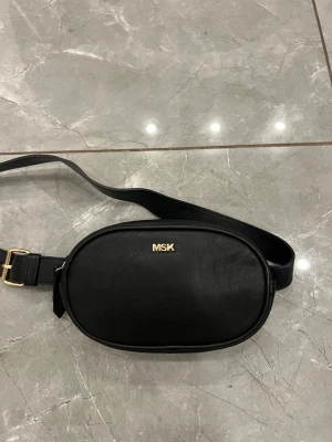 Svart midjeväska från MSK i skinn - Säljer en stilren svart midjeväska från MSK med justerbart band och guldfärgade detaljer. Väskan är ovalformad och har en dragkedja upptill samt flera fack inuti för småsaker. Det går även att ta bort bandet och använda den som en handväska.
