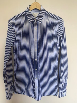 Blåvit randig skjorta Oscar Jacobson -  Snygg randig skjorta från Oscar Jacobson, storlek 38. 100% bomull. Skjortan har en klassisk krage med en elegant rund skjortnål,en snygg detalj som ger kragen ett unikt och stiligt intryck.