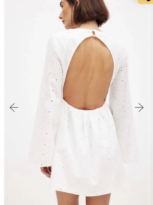 Studentklänning  - Superfin vit kort klänning med broderade blommor, djup v-ringning och öppen rygg. Klänningen har långa ärmar och är gjord i bomullstyg med broderade detaljer. Perfekt för studenten!