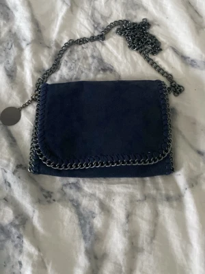 Mörkblå mocka axelväska med kedja - Snygg mörkblå axelväska i mocka med grov silverfärgad kedja som axelrem och dekorativ kedjedetalj runt kanten. Väskan har ett lock och en rund metallbricka vid kedjan. Perfekt för dig som gillar stilrena accessoarer med lite edge.