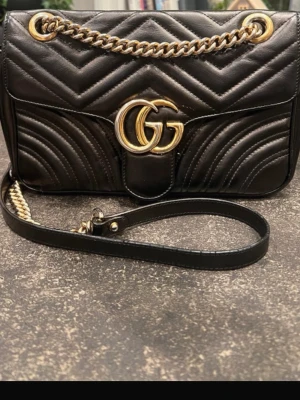 Svart quiltad axelväska från Gucci - Säljer en svart axelväska från Gucci i quiltat skinn med guldfärgade kedjedetaljer och GG-logga framtill. Väskan har ett hjärtmönster på baksidan och dragkedja inuti. Snygg och stilren design som lyfter din look.
