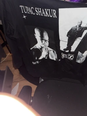 Svart Tupac Shakur långärmad tröja - Säljer en svart långärmad tröja med stort Tupac Shakur-tryck framtill. Tröjan har två stora svartvita fotoprints och texten 'Tupac Shakur' upptill. Perfekt för dig som gillar streetwear och hiphopstil.