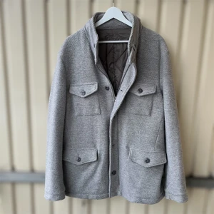 Grå ulljacka från Gran Sasso - Snygg grå jacka från Gran Sasso i mjuk ullmix. Jackan har fyra fickor med lock och knappar framtill, quiltat foder och både dragkedja och knäppning. Perfekt för dig som gillar stilren och klassisk look med modern touch. En knapp saknas men finns extra!