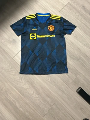 Manchester United bortatröja Adidas - Säljer en Manchester United bortatröja från Adidas med blå och svarta geometriska mönster, gula detaljer och klubbmärke på bröstet. Tröjan har TeamViewer-logga och klassiska Adidas-ränder på axlarna. Perfekt för dig som älskar fotboll och vill sticka ut på läktaren.