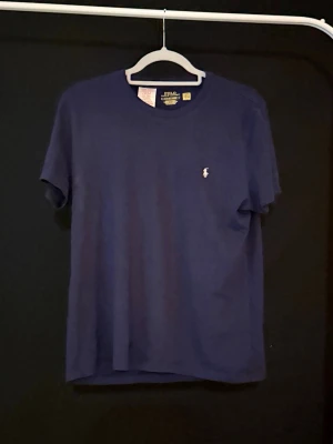 Marinblå t-shirt från Polo Ralph Lauren - Snygg marinblå t-shirt från Polo Ralph Lauren med klassisk vit logga broderad på bröstet. Modellen är kortärmad och har en rund hals. Materialet är mjuk bomull som känns skönt mot huden. Passformen är normal och funkar perfekt till jeans eller shorts.