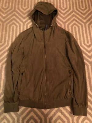 Olivgrön vindjacka med camo-detaljer - Snygg olivgrön vindjacka från Zara Man med huva och hel dragkedja. Insidan har coola camouflage-detaljer och jackan har två fickor med dragkedja. Perfekt för dig som gillar streetstyle och vill ha en lätt jacka med skön passform.