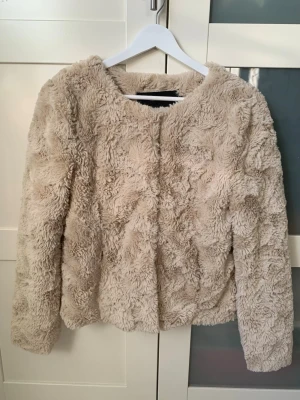 Beige jacka/kavaj från Vero Moda - Supermjuk och fluffig jacka/kavaj i beige från Vero Moda. Jackan har rund hals, långa ärmar och knäppning med knappar framtill. Insidan är fodrad med ljus polyester.