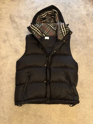 Svart pufferjacka från Burberry - Säljer en svart pufferjacka från Burberry med avtagbara ärmar så den kan användas som väst. Jackan har huva med klassiskt rutigt foder i grått, vitt och rött. Två dragkedjefickor fram och Burberry-patch på ärmen. Perfekt för kalla dagar. Skriv vid minsta fråga eller fundering