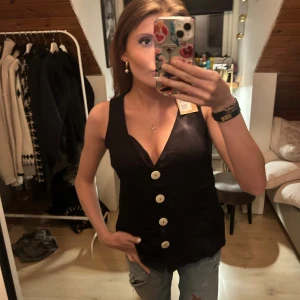 Svart väst med stora knappar - Säljer en svart väst med djup v-ringning och fyra stora vita knappar framtill. Västen har en figurnära passform och är ärmlös, perfekt att styla över en t-shirt eller skjorta. Materialet ser ut att vara slätt och lite glansigt.