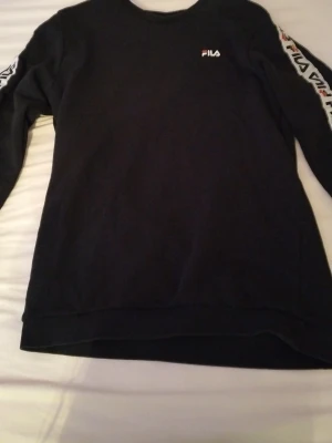 Svart långärmad tröja från FILA - Svart långärmad tröja från FILA i storlek M. Tröjan har logga på bröstet och tryck längs ärmarna. Klassisk rund halsringning och normal passform. Perfekt för en sportig och avslappnad stil.