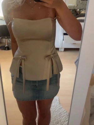 Guldig satin korsett med rosetter - Säljer en guldig korsettopp i satin med axelbandslös design. Toppen har två dekorativa rosetter framtill och en smidig dragkedja i ryggen. Snygg, figurnära passform som ger en stilren siluett. Perfekt att styla med jeans eller kjol för en trendig look.