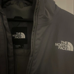 Grå vindjacka från The North Face - Säljer en stilren grå vindjacka från The North Face med huva och dragkedja framtill. Jackan har en enkel design med logga på bröstet och är perfekt för blåsiga dagar. Passar dig som gillar funktionella och snygga ytterplagg. Funkar också för regnigt väder. Köpt för 1900kr