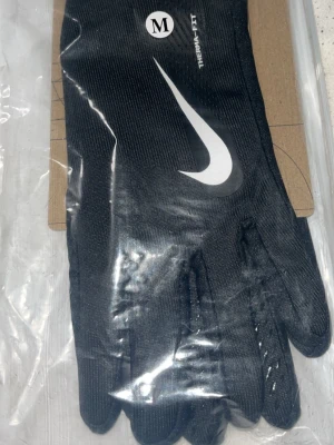 Svarta Nike Therma-Fit handskar - Svarta Nike Therma-Fit handskar med vit swoosh-logga på ovansidan och greppdetaljer i handflatan. Handskarna är tillverkade i syntetmaterial och har en elastisk mudd för extra komfort. Perfekta för träning utomhus under kyliga dagar.