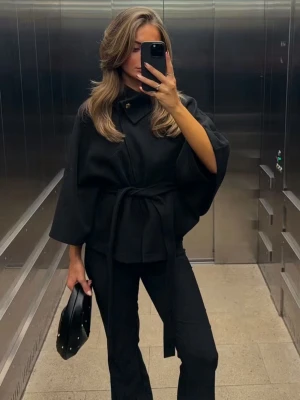 Svart cape kappa - Stilren svart cape-kappa med vida ärmar och knytskärp i midjan. Hög krage med knappstängning framtill. Perfekt för dig som vill ha en trendig och elegant look. Väldigt skön och passar alla outfits.