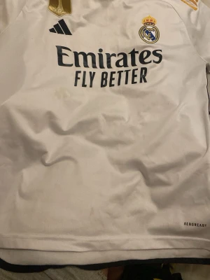 Real Madrid matchtröja Adidas vit - Säljer en vit Real Madrid matchtröja från Adidas med klubbmärke, svart Adidas-logga och guldmärke på bröstet. Tröjan har svart text 'Emirates Fly Better' och är tillverkad i lätt och ventilerande AEROREADY-material.