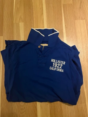 Blå pikétröja från Hollister - Mörkblå pikétröja från Hollister med vit text 'Hollister 1922 California' på bröstet. Klassisk krage med vit kant och knappar framtill. Perfekt till jeans eller shorts för en avslappnad look.