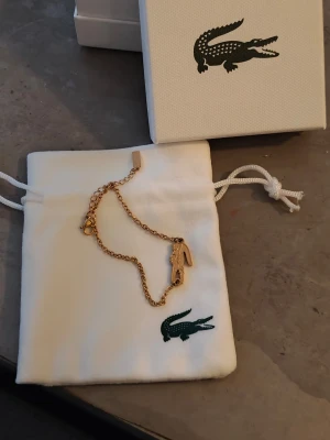 Guldigt armband från Lacoste - Snyggt guldigt armband från Lacoste med den klassiska krokodilberlocken. Kedjan är tunn och elegant, perfekt för att addera lite stil till din outfit. Kommer i originalpåse och ask med Lacoste-logga.