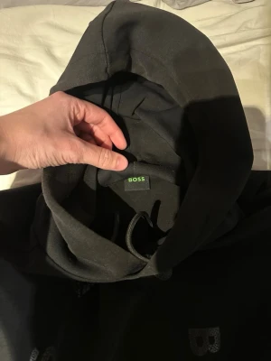 Svart hoodie från BOSS - Säljer en svart hoodie från BOSS med klassisk huva och dragsko. Hoodien har diskreta BOSS-loggor framtill och är tillverkad i mjukt bomullsmaterial för en skön och avslappnad look. Perfekt för dig som gillar stilrena och tidlösa plagg. Kan diskutera pris