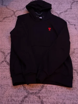 Svart hoodie från Ami Paris med rött hjärta - Säljer en svart hoodie från Ami Paris med klassisk huva och dragsko. På bröstet finns Ami:s ikoniska röda hjärtemblem. Hoodien har en känguruficka framtill och ribbade muddar vid ärmar och nederkant. Perfekt för en avslappnad och stilren look.