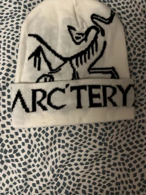 Vit mössa från Arc'teryx - Snygg vit mössa från Arc'teryx med svart logga och text framtill. Mössan har uppvikt kant och är tillverkad i mjukt material som passar perfekt för kalla dagar. Den har en modern och sportig look med tydlig branding.