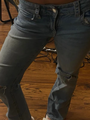 Blå slitna straight jeans - Säljer ett par blå jeans med slitningar och hål på knäna. Jeansen har en rak passform och klassisk femficksdesign. Perfekta för en avslappnad och trendig look.