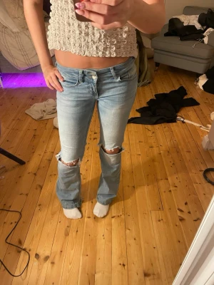 Ljusblå bootcut jeans med slitningar - Säljer ett par ljusblå bootcut jeans med slitna knän och fransiga detaljer. Jeansen har låg midja och klassisk femficksdesign. Perfekta för en avslappnad och trendig look. 