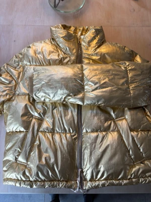 Guldig pufferjacka med dragkedja Coster Copenhagen - Snygg och trendig pufferjacka i metallic guld med quiltad design och hög krage. Jackan har en något kortare passform, långa ärmar och dragkedja framtill. Väldigt fint foder. Perfekt för dig som vill sticka ut och hålla värmen under kalla dagar. Nypris 1799kr