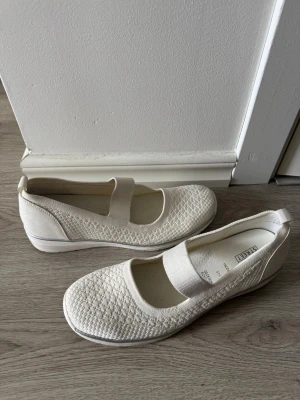 Vita ballerinaskor med resårband - Snygga vita ballerinaskor från Skechers med stickad ovandel och elastiskt band över foten för extra komfort. Skorna har rund tå och platt sula, perfekta för en avslappnad och stilren look. Materialet är mjukt och andas, vilket gör dem sköna att bära hela dagen.