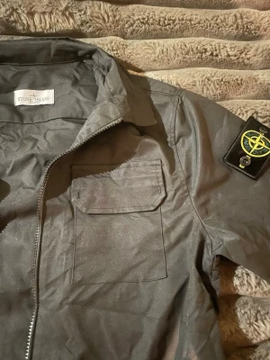 Svart overshirt från Stone Island - Snygg svart overshirt från Stone Island med klassisk patch på ärmen. Jackan har dragkedja framtill, bröstficka med lock och krage. Perfekt för lager-på-lager och streetwear-stil.