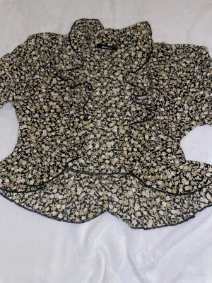 Blommig volangblus/kofta från Gina Tricot - Söt blus med svart botten och små blommor i beige, gul, vitt och ljusrosa. Volangdetaljer längs fram och vid kragen ger en romantisk vibe. 