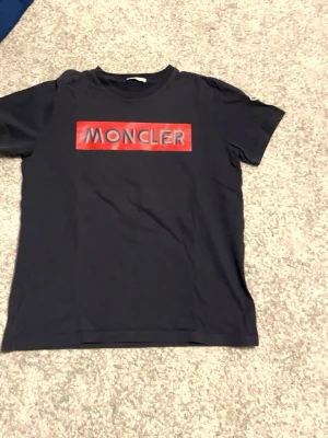 Svart Moncler t-shirt med röd logga - Snygg svart t-shirt från Moncler med stor röd logga på bröstet och ett broderat Moncler-märke på ärmen. Klassisk passform och rund halsringning. Perfekt för dig som vill ha en stilren och trendig look.