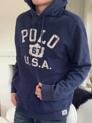 Marinblå hoodie från Polo Ralph Lauren - Säljer en marinblå hoodie från Polo Ralph Lauren med stor vit vintage-inspirerad text 'POLO 67 U.S.A.' på bröstet. Hoodien har huva med dragsko, magficka och ribbade muddar. Perfekt för en avslappnad och sportig stil.