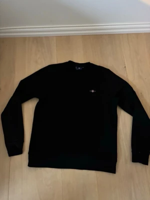 Svart sweatshirt från GANT - Klassisk svart sweatshirt från GANT med rund hals och diskret logga på bröstet. Tröjan har lång ärm och är tillverkad i mjukt material som passar perfekt till vardagslooken.
