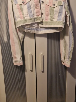 Pastellrandig croppad jeansjacka - Säljer en croppad jeansjacka med vertikala pastellränder i rosa, blått, grönt och vitt. Jackan har klassisk knappstängning framtill, två bröstfickor med lock och silverfärgade knappar. Perfekt för dig som gillar färg och vill sticka ut. Inte använd, prislappen är inte kvar. Vi har katt i hemmet.