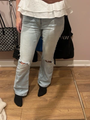 Ljusblå bootcut jeans med slitningar - Säljer ett par ljusblå jeans med bootcut passform och slitna hål över knäna. Jeansen har klassisk femficksdesign, hög midja och är tillverkade i ett mjukt denimtyg. Perfekta för en avslappnad och trendig look.