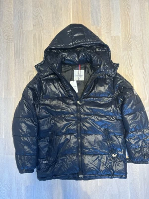 Svart dunjacka från Moncler - Snygg svart dunjacka från Moncler med glansig finish och huva. Jackan har quiltad design, dragkedja framtill och två sidofickor med dragkedja. Perfekt för kalla dagar och ger en cool streetwear-look. Äkthets verifierad av winted! 