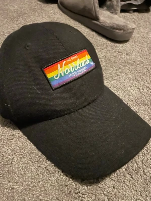 Svart keps Norrland Satin & Company - Svart keps med böjd skärm och broderad patch framtill med regnbågsfärger och texten 'The Great Norrland'. Justerbar rem bak med metallspänne och broderad logga 'Satin & Company'. Snygg och stilren sopparkeps i bomull.