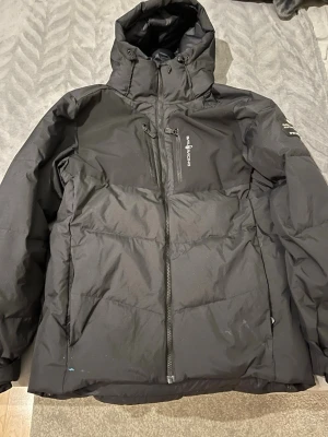 Svart dunjacka från sail racing  - Säljer en svart dunjacka från sail racing med huva och dragkedja framtill. Jackan har en bröstficka med dragkedja och logga på bröstet. Perfekt för kalla dagar och har en klassisk pufferstil som håller dig varm.