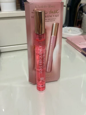 Charlotte Tilbury Love Frequency parfym - En snygg parfym från Charlotte Tilbury i en smal, cylinderformad rosa glasflaska med guldfärgat lock. Flaskan har texten 'Love Frequency' och en liten blom-ikon. Kommer i en matchande rosa förpackning.