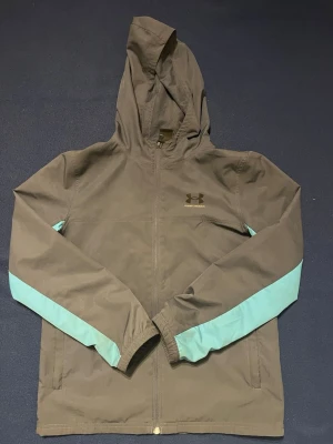 Grå vindjacka från Under Armour - Snygg grå vindjacka från Under Armour med turkosa detaljer längs ärmarna och huva. Jackan har dragkedja framtill, två sidofickor och elastiska muddar vid ärmsluten. Perfekt för blåsiga dagar och sportiga outfits. Har en liten fläck på magen.