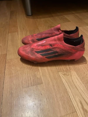 Röda adidas fotbollsskor utan snörning - Säljer ett par röda adidas fotbollsskor med svarta detaljer och utan snörning. Skorna har en strömlinjeformad design och är tillverkade i syntetmaterial för bästa prestanda på planen. Perfekta för dig som vill ha stilrena och snabba skor till matchen.
