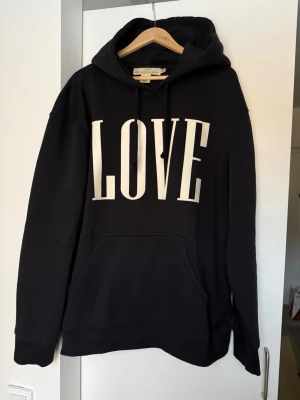 Svart hoodie med LOVE-tryck - Svart hoodie med stort vitt LOVE-tryck på bröstet. Klassisk känguruficka framtill, justerbar huva med snören och ribbade muddar. Tillverkad i mjukt bomullsmaterial som är skönt att bära.