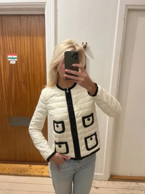 Vit Moncler jacka med svarta detaljer - Snygg vit Moncler dunjacka med svarta kontrastkanter och fyra dekorativa fickor framtill. Jackan har rund halsringning, lång ärm och stängs med dragkedja. Nätt och fin. 